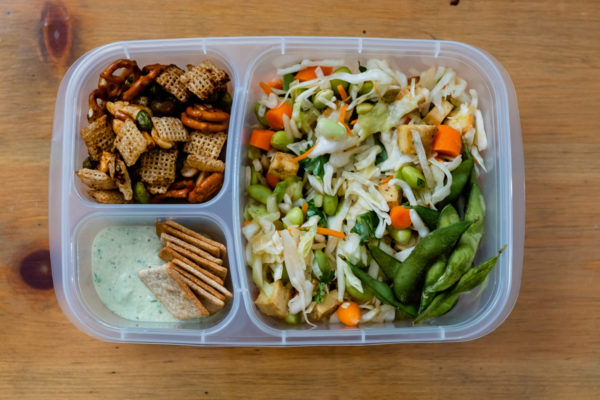 A meal prep container is filled with an assortment of soy foods like a salad and crackers with dip.