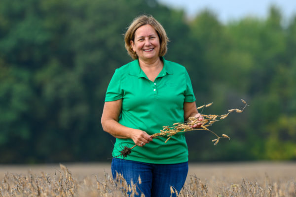 Laurie Isley smiles and stands in a soy field while holding a soybean stalk.