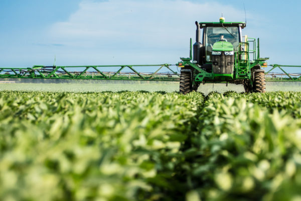 A green tractor drives a crop sprayer over a soybean field.