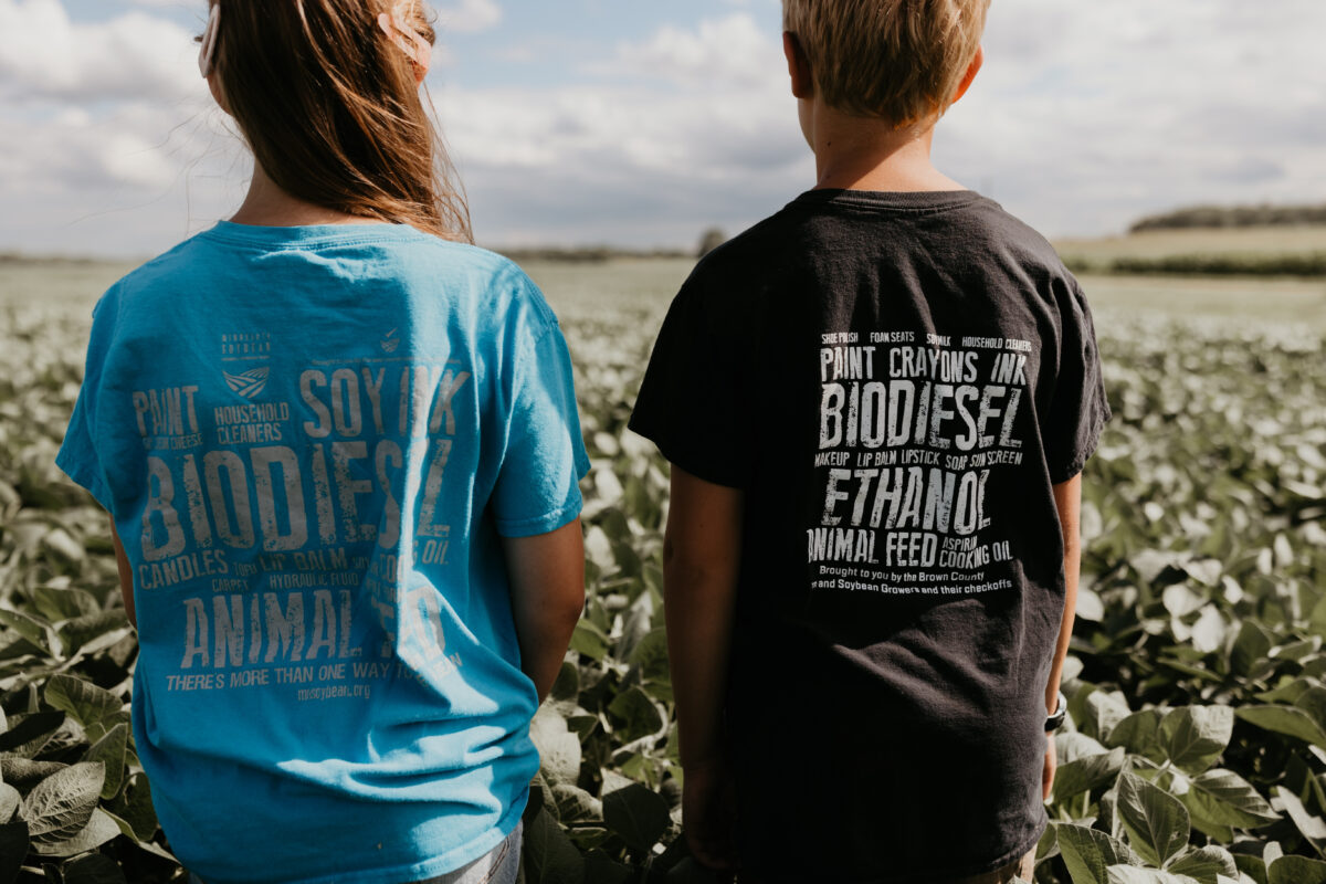 A girl and boy standing next to each other in a field of soybeans with graphic t-shirts on that highlight the many uses of soy.